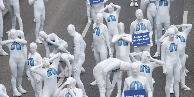 Demonstranten stehen in weißen Ganzkörperanzügen und blau aufgedruckten Lungensymbolen auf einer Straße Demonstranten stehen in weißen Ganzkörperanzügen und blau aufgedruckten Lungensymbolen auf einer Straße