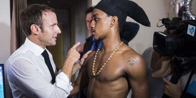 Macron spricht mit einem Mann mit Kopftuch und freiem Oberkörper. Er zeigt mahnend mit dem Zeigefinger auf seinen Gesrpächspartner. Macron spricht mit einem Mann mit Kopftuch und freiem Oberkörper. Er zeigt mahnend mit dem Zeigefinger auf seinen Gesrpächspartner.