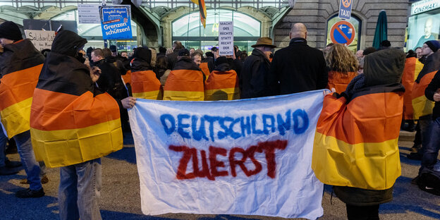 Menschen in schwarz-rot-gelbe Fahnen gehüllt, halten Transparent hoch. Darauf steht: "Deutschland zuerst" Menschen in schwarz-rot-gelbe Fahnen gehüllt, halten Transparent hoch. Darauf steht: "Deutschland zuerst"