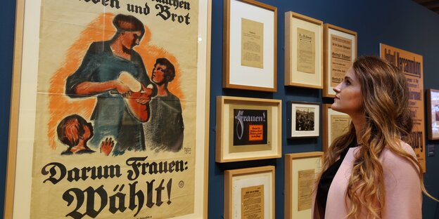 Eine Frau blickt auf historische Plakate zum Frauenwahlrecht, auf einem steht "Eure Kinder brauchen Frieden und Brot. Darum Frauen: Wählt!"