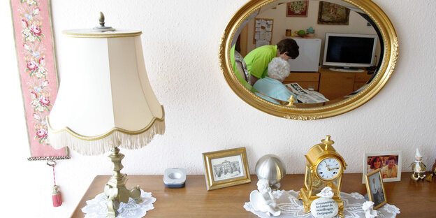 Kommode mit einer Lampe und einem Spiegel, in dem eine Pflegerin zu sehen ist Kommode mit einer Lampe und einem Spiegel, in dem eine Pflegerin zu sehen ist