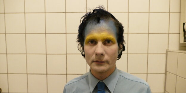 Ein Mann mit blauer und goldener Farbe auf der Stirn steht in einem Badezimmer Ein Mann mit blauer und goldener Farbe auf der Stirn steht in einem Badezimmer