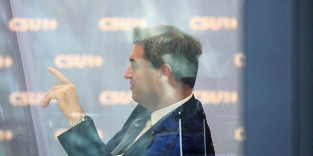 Markus Söder hinter einer Glasscheibe und hebt einen Finger Markus Söder hinter einer Glasscheibe und hebt einen Finger
