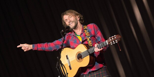 Ein Mann im Karohemd und mit Gitarre zeigt nach rechts