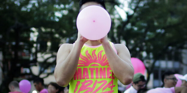 Ein Mensch in einem gelb-pinken Tanktop bläst einen rosa Luftballon auf, sein Gesicht wird dadurch verdeckt Ein Mensch in einem gelb-pinken Tanktop bläst einen rosa Luftballon auf, sein Gesicht wird dadurch verdeckt