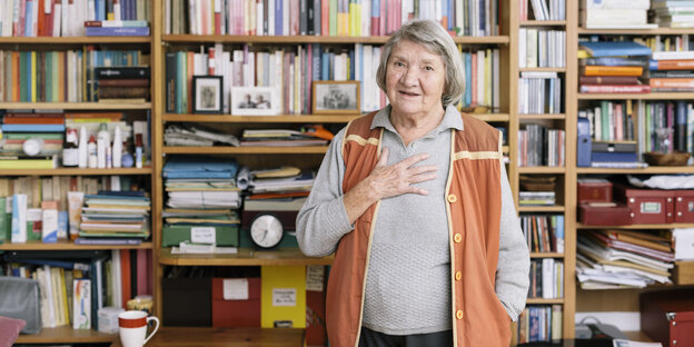 Eine ältere Frau steht vor einem Bücherregal und hält sich eine Hand vor die Brust Eine ältere Frau steht vor einem Bücherregal und hält sich eine Hand vor die Brust