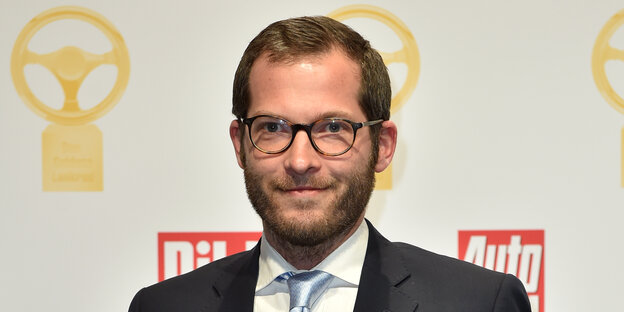 Ein Mann im Anzug steht vor einer weißen Wand auf der mehrere Logos abgebildet sind. Ein Mann im Anzug steht vor einer weißen Wand auf der mehrere Logos abgebildet sind.