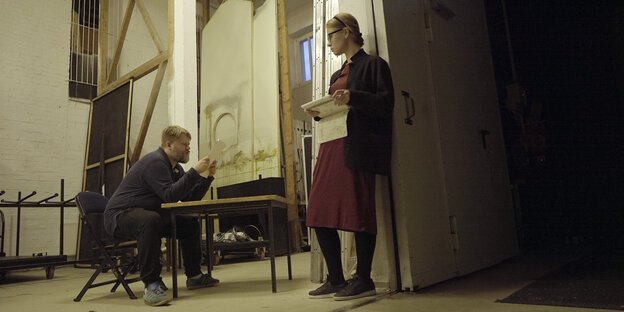 Der Regisseur sitzt gebeugt an einem Tisch, an der Wand lehnt seine Frau. Der Regisseur sitzt gebeugt an einem Tisch, an der Wand lehnt seine Frau.