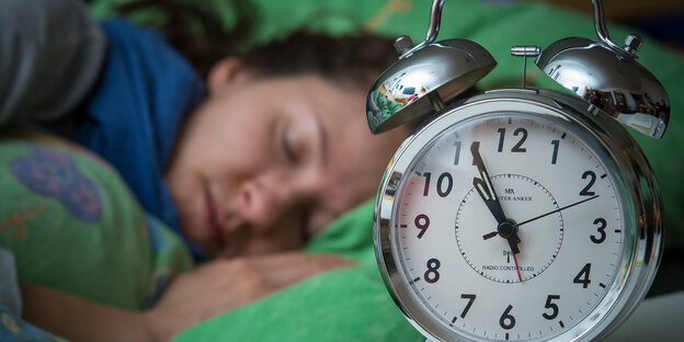 Eine Frau schläft im Bett, neben ihr auf dem Nachttischränkchen steht ein Wecker. Eine Frau schläft im Bett, neben ihr auf dem Nachttischränkchen steht ein Wecker.