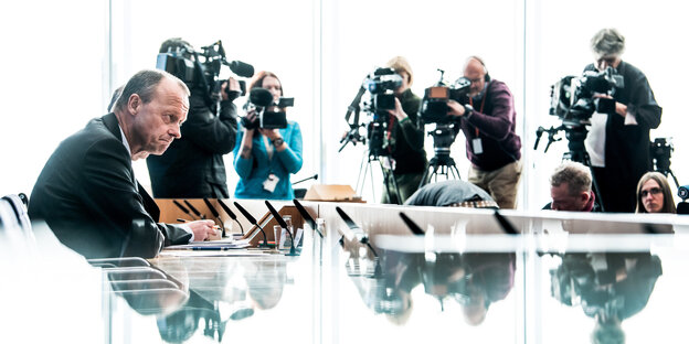 Friedrich Merz sitzt an einem Tisch, dahinter Fotografen Friedrich Merz sitzt an einem Tisch, dahinter Fotografen
