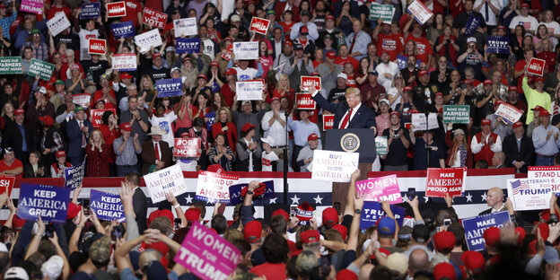 Viele Menschen in einer Halle schwenken Plakate, mittendrin steht US-Präsident Donald Trump vor einem Rednerpult.