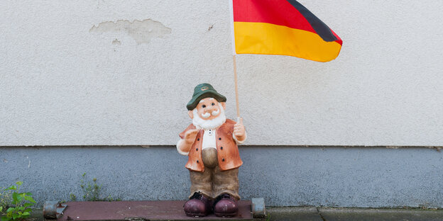 Ein Gartenzwerg steht vor einer Hauswand, in der Hand hält er eine Deutschlandflagge Ein Gartenzwerg steht vor einer Hauswand, in der Hand hält er eine Deutschlandflagge