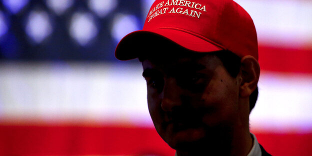 Ein Mann steht vor einer amerikanischen Flagge. Auf seinem Kopf trägt er eine Cap mit dem Spruch: Make America great again Ein Mann steht vor einer amerikanischen Flagge. Auf seinem Kopf trägt er eine Cap mit dem Spruch: Make America great again