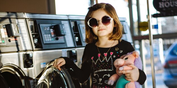 Ein Mädchen mit großer Sonnenbrille und Kuscheltier in der Hand steht an einer Waschmaschine Ein Mädchen mit großer Sonnenbrille und Kuscheltier in der Hand steht an einer Waschmaschine