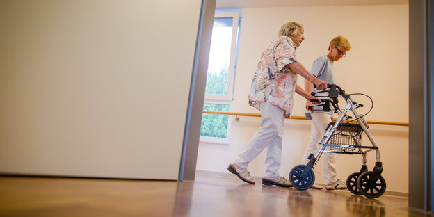 Eine Pflegekraft und eine Frau mit Rollator gehen über einen Flur.