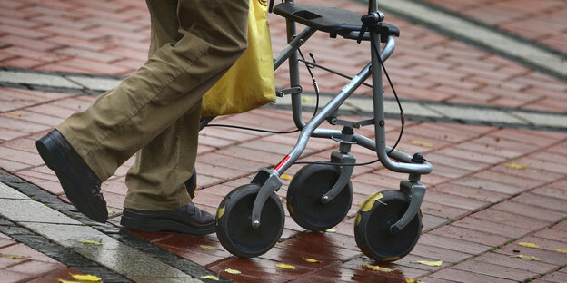 Die Beine eines Mannes sind zu sehen, der auf einer nassen Straße mit einem Rollator läuft Die Beine eines Mannes sind zu sehen, der auf einer nassen Straße mit einem Rollator läuft