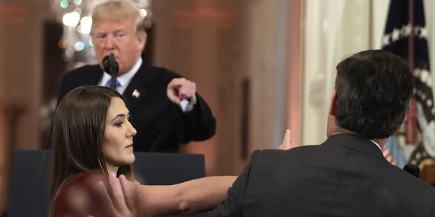 Donald Trump hält eine Rede an einem Rednerpult und zeigt mit dem Finger auf einen Mann vor ihm. Dem Mann wird von einer Frau sein Mikrofon weggenommen Donald Trump hält eine Rede an einem Rednerpult und zeigt mit dem Finger auf einen Mann vor ihm. Dem Mann wird von einer Frau sein Mikrofon weggenommen