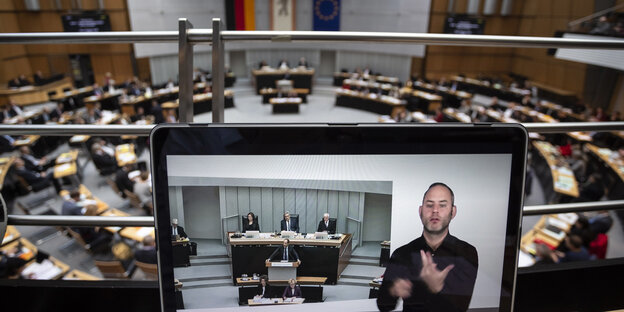 Auf einem Bildschirm ist ein Mann zu sehen, der in Gebärdensprache spricht. Im Hintergrund ist der Plenarsaal des Berliner Abgeordnetenhauses zu sehen Auf einem Bildschirm ist ein Mann zu sehen, der in Gebärdensprache spricht. Im Hintergrund ist der Plenarsaal des Berliner Abgeordnetenhauses zu sehen