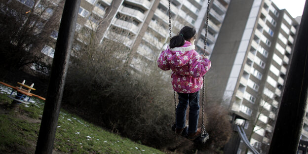Ein Mädchen in pinker Jacke Schaukelt vor einer Kulisse aus Plattenbauten. Ein Mädchen in pinker Jacke Schaukelt vor einer Kulisse aus Plattenbauten.