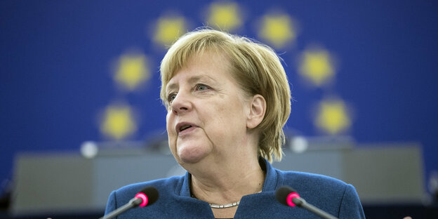 Merkel steht am Rednerpult, im Hintergrund ist die EU-Flagge. Die Sterne stehen über ihrem Kopf und sehen aus wie ein Heiligenschein Merkel steht am Rednerpult, im Hintergrund ist die EU-Flagge. Die Sterne stehen über ihrem Kopf und sehen aus wie ein Heiligenschein