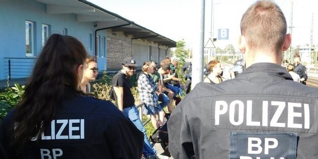 Zwei Polizisten von hinten, dahinter lehnen Menschen an einem Zaun, ihre Gesichter sind unkenntlich gemacht. Zwei Polizisten von hinten, dahinter lehnen Menschen an einem Zaun, ihre Gesichter sind unkenntlich gemacht.