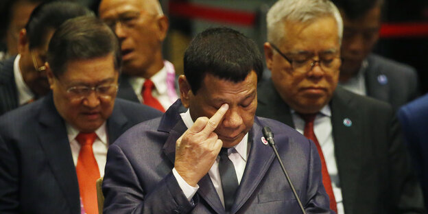 Der philippinische Präsident Rodrigo Duterte hält seinen Mittelfinger an sein Auge und sitzt zwischen zwei Personen Der philippinische Präsident Rodrigo Duterte hält seinen Mittelfinger an sein Auge und sitzt zwischen zwei Personen