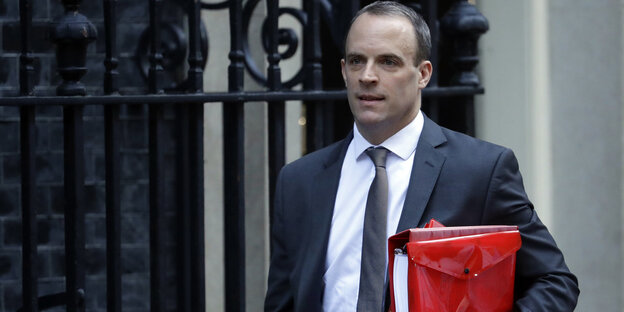 Dominic Raab läuft mit roter Tasche vor einem Zaun lang Dominic Raab läuft mit roter Tasche vor einem Zaun lang