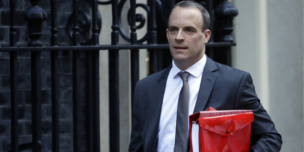 Dominic Raab läuft mit roter Tasche vor einem Zaun lang