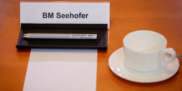 Auf einem Namenschild steht BM Seehofer. Davor liegen ein weißes DIN A4 Blatt sowie zwei Bleistifte. Daneben steht eine weiße Porzellantasse mit Untersetzer Auf einem Namenschild steht BM Seehofer. Davor liegen ein weißes DIN A4 Blatt sowie zwei Bleistifte. Daneben steht eine weiße Porzellantasse mit Untersetzer