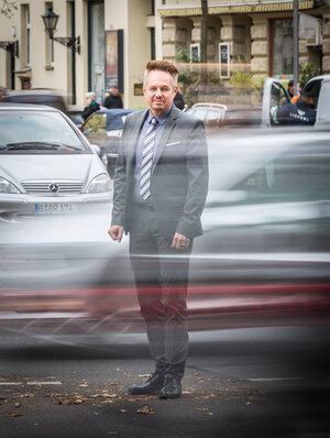 Ein Mann in Anzug steht auf einer Straße, hinter ihm fährt ein Auto vorbei Ein Mann in Anzug steht auf einer Straße, hinter ihm fährt ein Auto vorbei