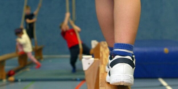 Ein Kinderbein auf einem Schwebebalken. In Entfernung Kinder, die versuchen ein Seil hochzuklettern. Seil Ein Kinderbein auf einem Schwebebalken. In Entfernung Kinder, die versuchen ein Seil hochzuklettern. Seil