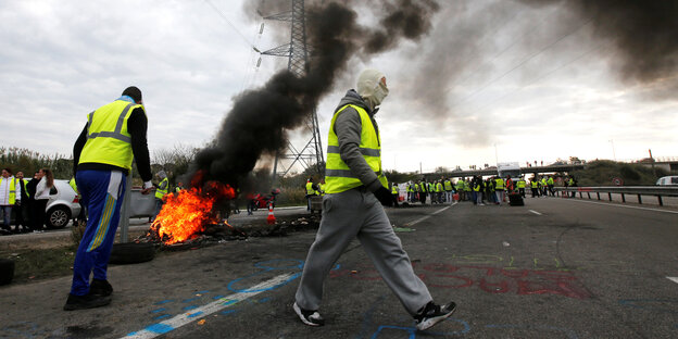 Zwei Protestler stehen in gelben Westen neben einer brennenden Barrikade Zwei Protestler stehen in gelben Westen neben einer brennenden Barrikade
