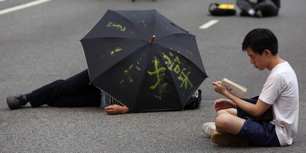 Ein Mensch sitzt auf der Straße, ein anderer liegt unter einem Regenschirm Ein Mensch sitzt auf der Straße, ein anderer liegt unter einem Regenschirm