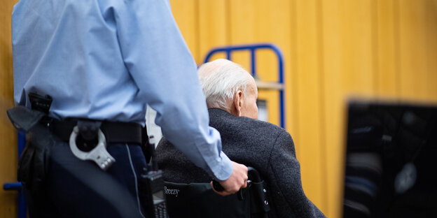 Ein Justizbeamter schiebt den Angeklagten, er sitzt im Rollstuhl, in den Gerichtssaal Ein Justizbeamter schiebt den Angeklagten, er sitzt im Rollstuhl, in den Gerichtssaal
