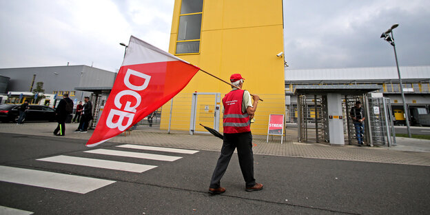 Ein Mann in einer roten Weste steht während eines Streiks bei Amazon mit einer DGB-Flagge vor dem Unternehmensstandort in Leipzig Ein Mann in einer roten Weste steht während eines Streiks bei Amazon mit einer DGB-Flagge vor dem Unternehmensstandort in Leipzig