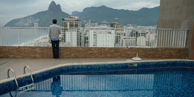 Ein Mann steht hinter einem Pool und guckt von der Veranda auf ein Stadtpanorama Ein Mann steht hinter einem Pool und guckt von der Veranda auf ein Stadtpanorama
