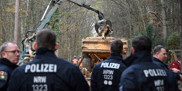 Ein Baggerarm hievt ein Baumstück auf einen Laster im Wald, im Vordergrund Rückenansichten von Polizisten