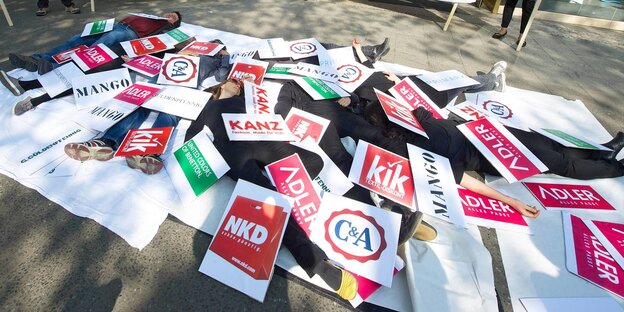 Demonstrant*innen liegen zwischen Schildern mit Namen von Modeketten Demonstrant*innen liegen zwischen Schildern mit Namen von Modeketten