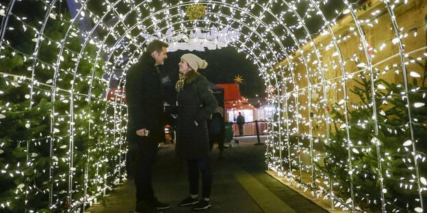 Ein Mann und eine Frau lächeln sich unter einem mit Lichterketten behängten Tunnel an Ein Mann und eine Frau lächeln sich unter einem mit Lichterketten behängten Tunnel an
