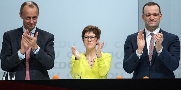 Zwei Männer in Anzügen und in der Mitte eine Frau in gelber Bluse applaudieren