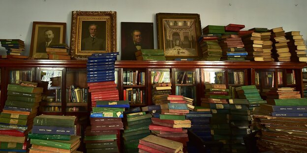In einer Bibliothek sind Bücherstapel und Porträts weißer Männer zu sehen In einer Bibliothek sind Bücherstapel und Porträts weißer Männer zu sehen