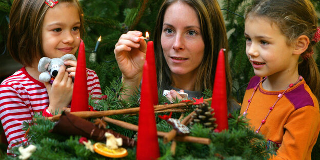 Eine Kita-Erzieherin zündet mit zwei Mädchen eine Kerze an einem Adventskranz an. Eine Kita-Erzieherin zündet mit zwei Mädchen eine Kerze an einem Adventskranz an.