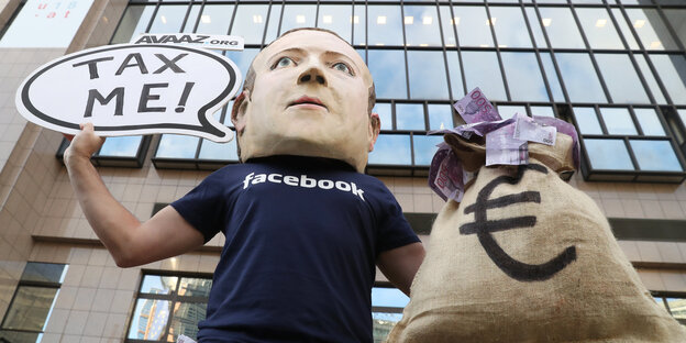 Ein Mann mit Mark-Zuckerberg-Maske hält ein Schild, auf dem "Tax me" steht Ein Mann mit Mark-Zuckerberg-Maske hält ein Schild, auf dem "Tax me" steht