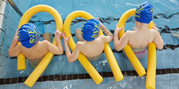 Drei Kinder sitzen am Beckenrad in Schwimmhosen und mit gelben Schwimmnudeln Drei Kinder sitzen am Beckenrad in Schwimmhosen und mit gelben Schwimmnudeln