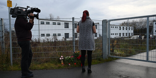 Ein Kameramann und eine Frau sind von hinten zu sehen, sie stehen am Zaun vor einer Unterkunft für Wohnungslose Ein Kameramann und eine Frau sind von hinten zu sehen, sie stehen am Zaun vor einer Unterkunft für Wohnungslose