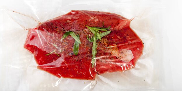 Ein in Plastikfolie verpacktes Stück Fleisch mit ein wenig frischen Gewürzen Ein in Plastikfolie verpacktes Stück Fleisch mit ein wenig frischen Gewürzen