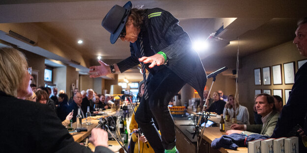 Udo Lindenberg, ein Mann in schwarzer Kleidung und mit schwarzem Hut tanzt auf einem Bartresen, viele Leute sitzen um ihn herum Udo Lindenberg, ein Mann in schwarzer Kleidung und mit schwarzem Hut tanzt auf einem Bartresen, viele Leute sitzen um ihn herum