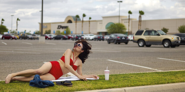 Eine Frau liegt im Bikini auf einem Grünstreifen auf dem Parkplatz eines Einkaufszentrums Eine Frau liegt im Bikini auf einem Grünstreifen auf dem Parkplatz eines Einkaufszentrums