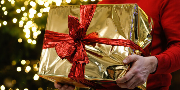 Ein Mann hält ein golden eingepacktes Weihnachtsgeschenk in den Händen. Im Hintergrund ist ein Weihnachtsbaum zu sehen. Ein Mann hält ein golden eingepacktes Weihnachtsgeschenk in den Händen. Im Hintergrund ist ein Weihnachtsbaum zu sehen.
