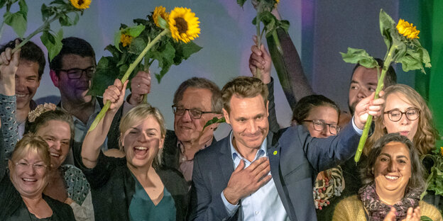 Mehrere Menschen winken mit Sonnenblumen von einer Bühne Mehrere Menschen winken mit Sonnenblumen von einer Bühne